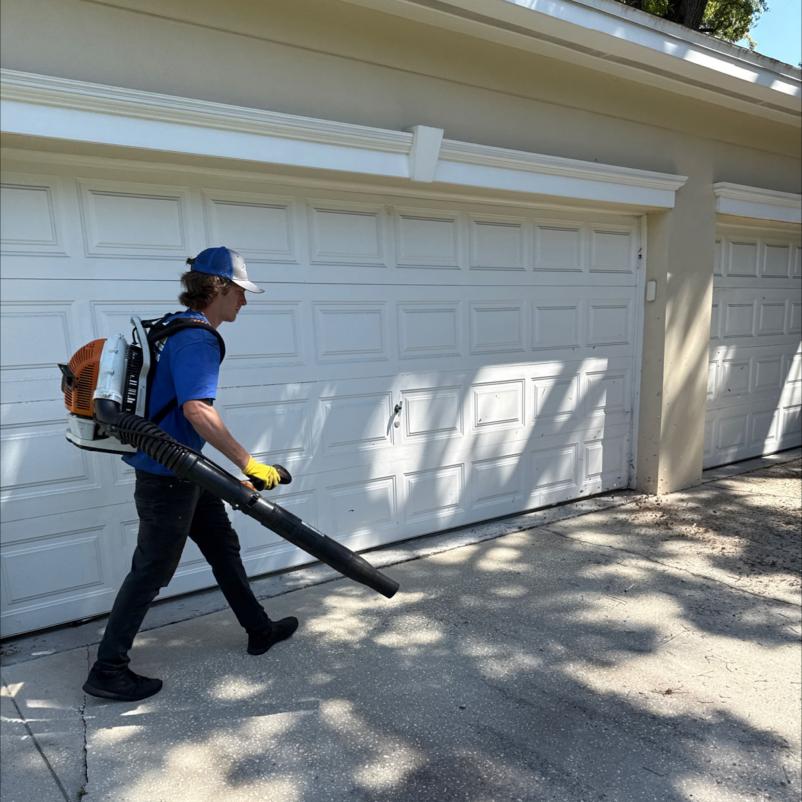 man using a backpack blower to clear debris from a driveway in front of a garage door